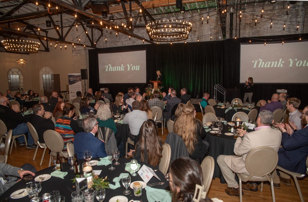 A large banquet hall is filled with people seated at round tables, watching a speaker on a stage. Two screens display the words Thank You. The room is decorated with string lights and chandeliers, creating a warm ambiance.