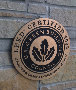 A round bronze plaque on a stone wall in Michigan displays the LEED Certified 2002 emblem from the U.S. Green Building Council, showcasing Leadership in Energy & Environmental Design 2.0 with a leaf design, symbolizing excellence in sustainable building.